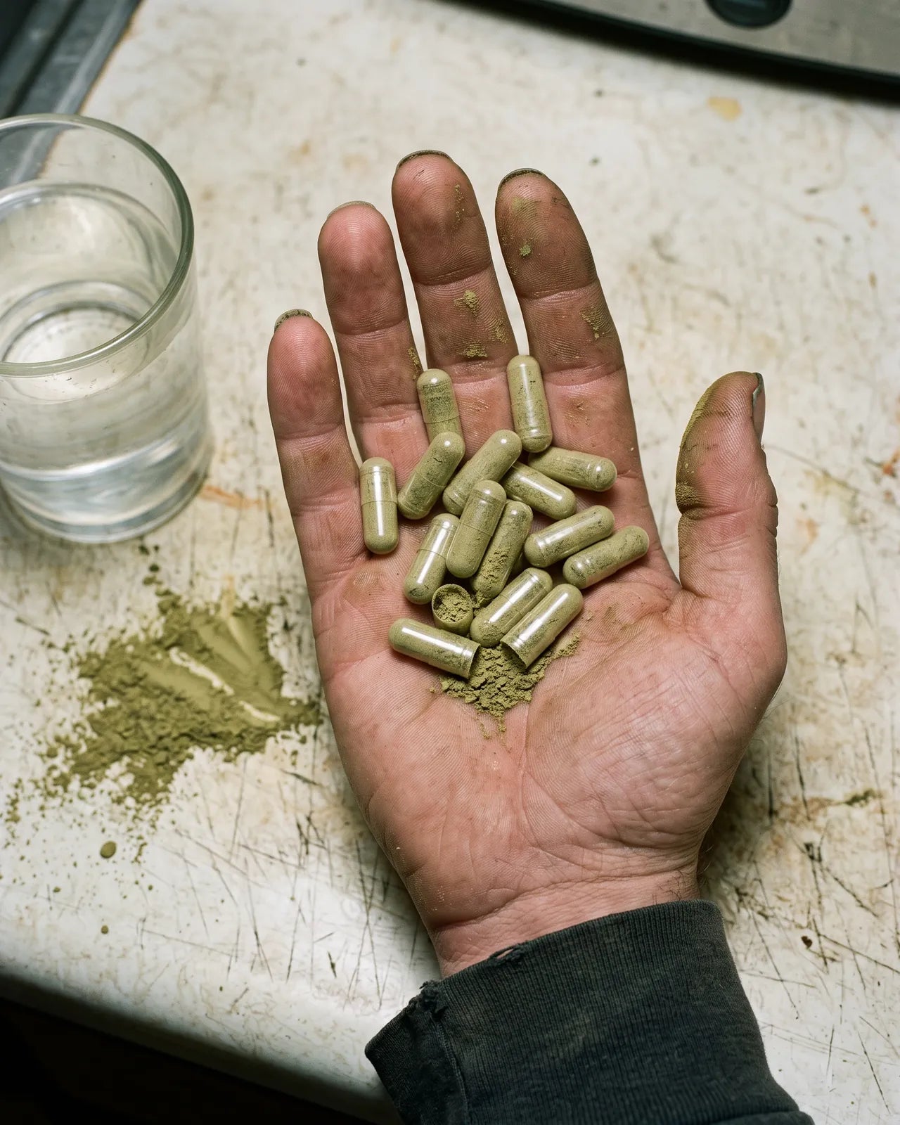 A grimy hand holding a pile of dusty green kratom capsules, one cracked open