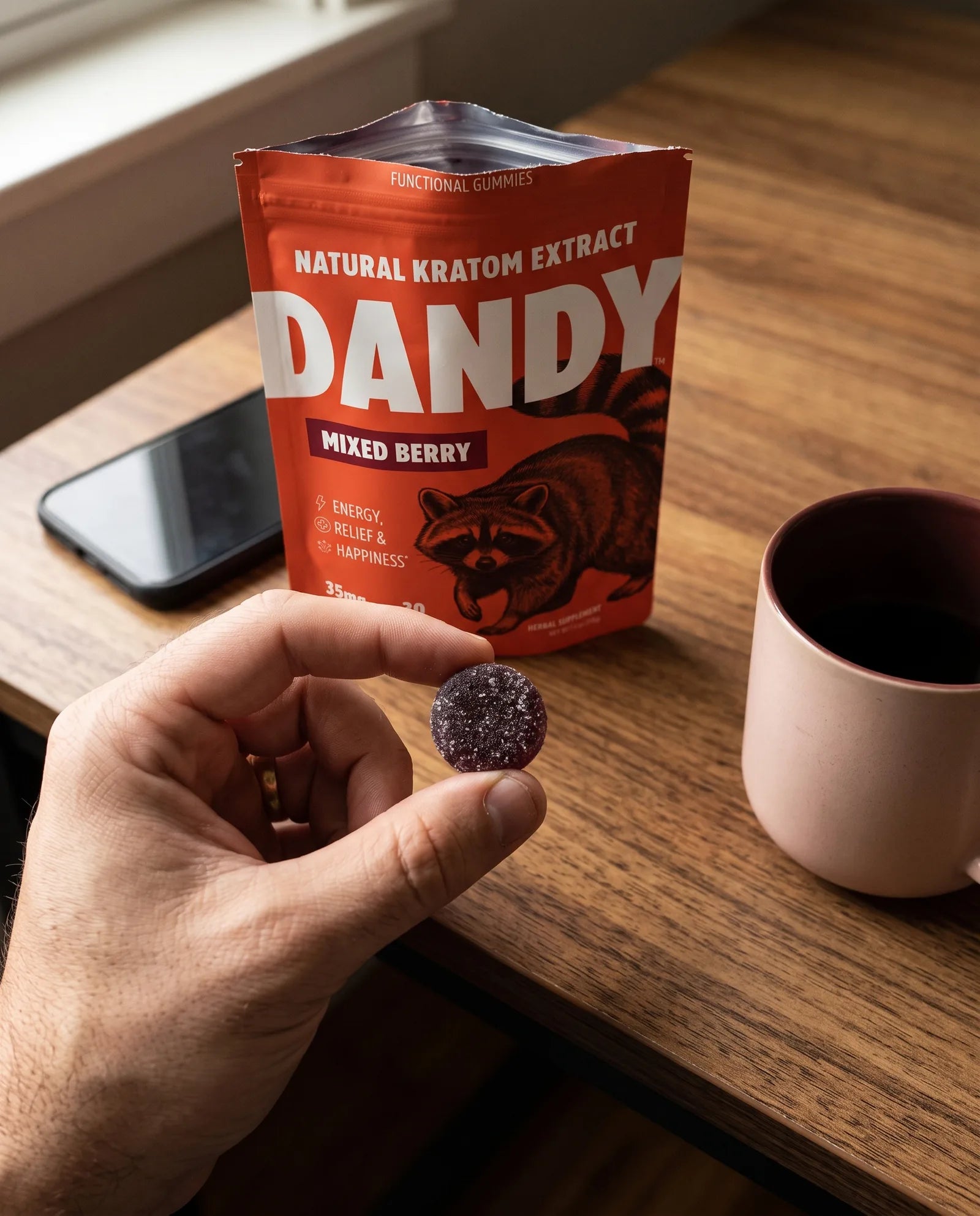 An adult holding a Dandy gummy over a desk with the open pouch beside a phone