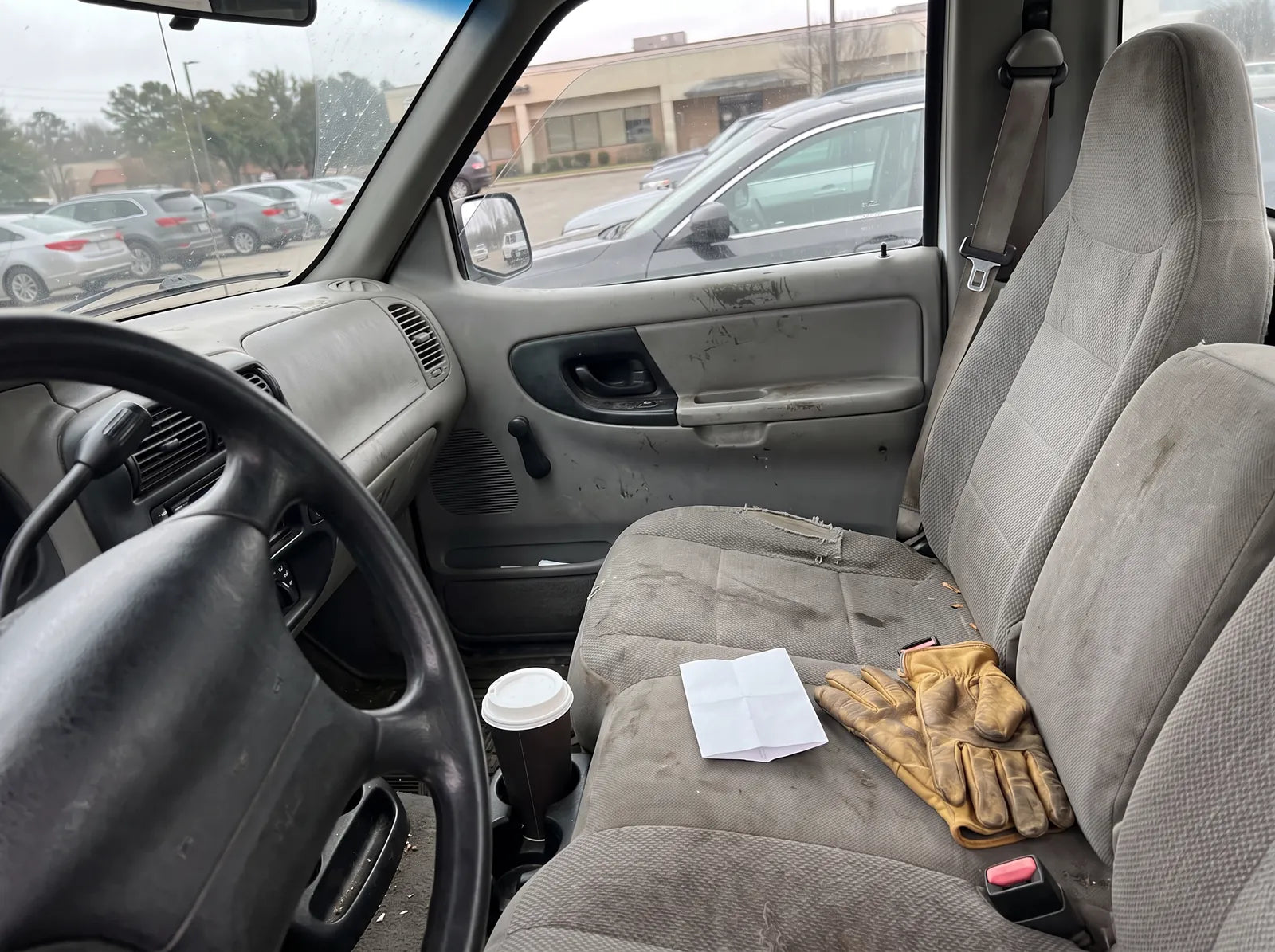 A folded prescription on a pickup truck’s passenger seat beside work gloves, a parking lot through the windshield