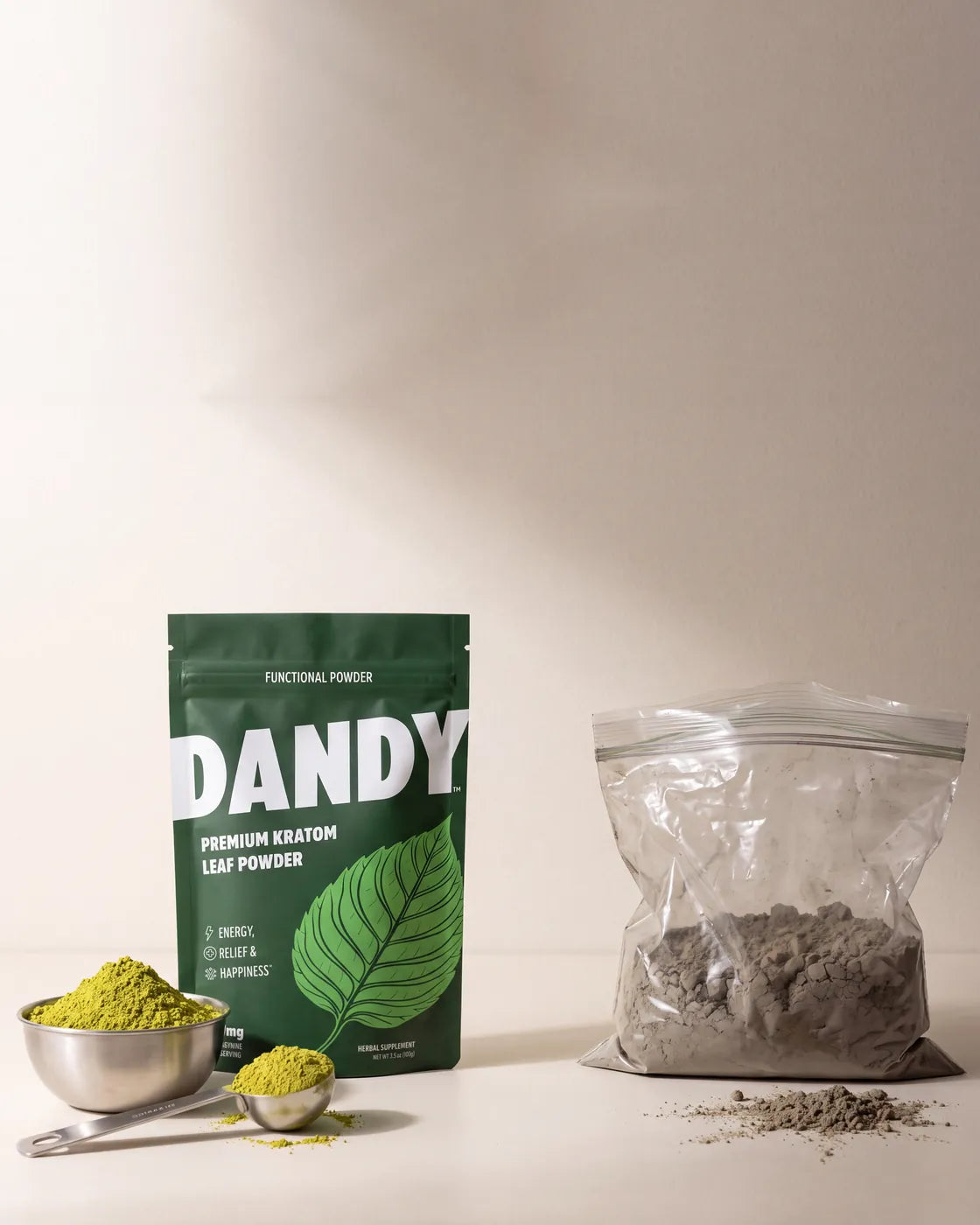 Fresh green raw leaf kratom powder beside the labeled Dandy pouch on one side, a plain unmarked bag of powder on the other.